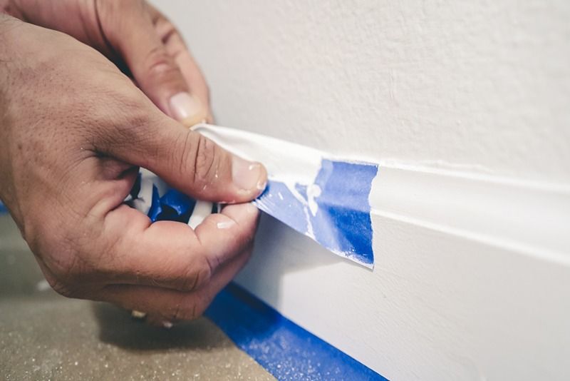 Removing_masking_tape_from_molding._A_painter_pulls_of_blue_painter's_tape_from_the_wall_to_reveal_a_clean_edge_baseboard.