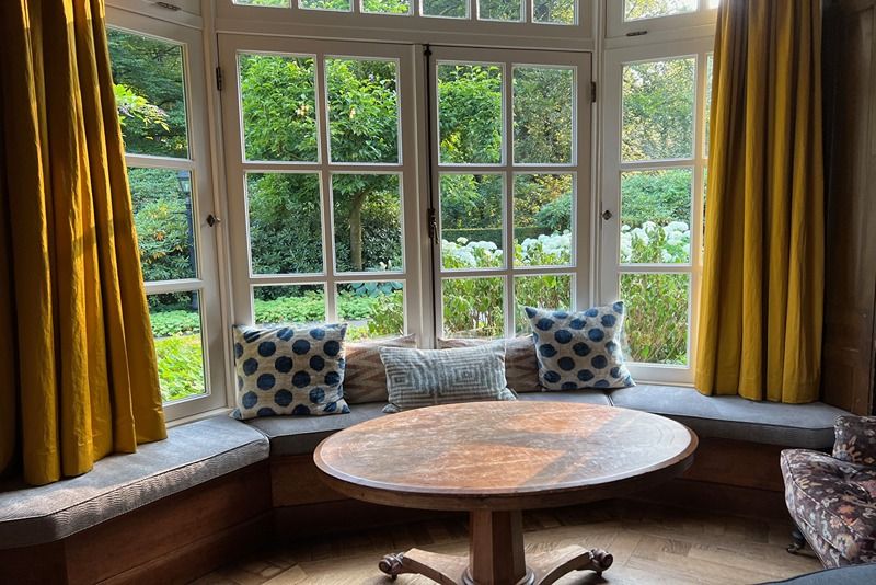Wooden_table_near_cozy_bay_window_seat_with_cushions_in_room._Interior_design