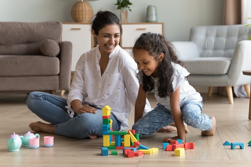 Indian_woman_and_daughter_play_wooden_blocks_seated_on_floor_smiling_looking_happy,_enjoy_carefree_weekend_leisure_and_pastime,_having_fun_together_at_modern_living_room._Playtime_at_home_with_child