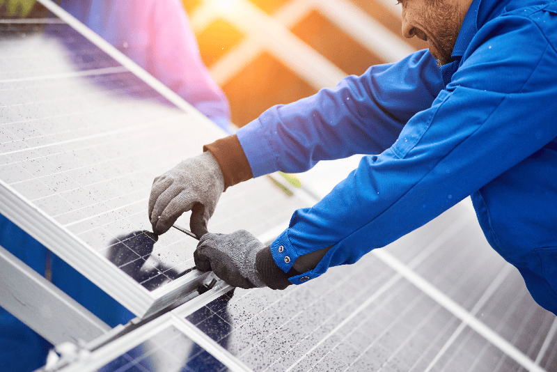 Smiling_male_technician_in_blue_suit_installing_photovoltaic_blue_solar_modules_with_screw._Man_electrician_panel_sun_sustainable_resources_renewable_energy_source_alternative_innovation